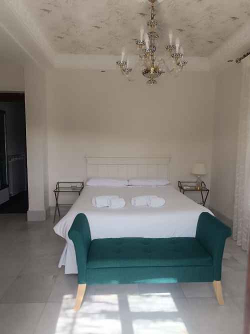 Dormitorio elegante en Suite Comares de Granada con araña, ropa de cama blanca, banco verde y pisos de azulejos