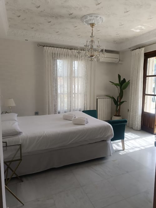 Habitación Bright Suite del hotel Comares en Granada con cama blanca, lámpara de araña, cortinas semitransparentes y vistas a la ciudad desde las puertas del balcón