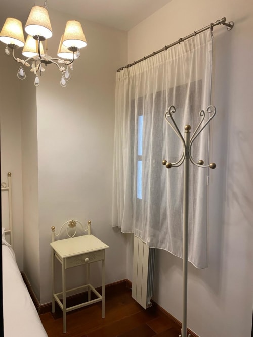 Elegante rincón del dormitorio en Suite Comares Granada con mesita de noche vintage, lámpara de araña, cortinas translúcidas y perchero decorativo