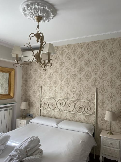 Elegante dormitorio en Suite Comares Granada con papel damasco, cama de hierro forjado, lámpara ornamental y ropa de cama blanca.