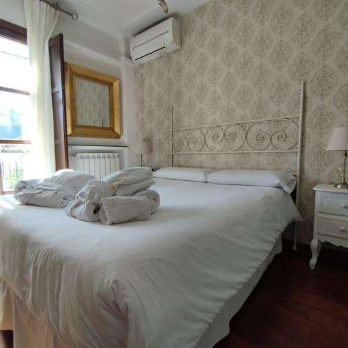 Dormitorio luminoso en Suite Comares Granada con cama de metal blanca, toallas enrolladas, papel tapiz damasco y aire acondicionado