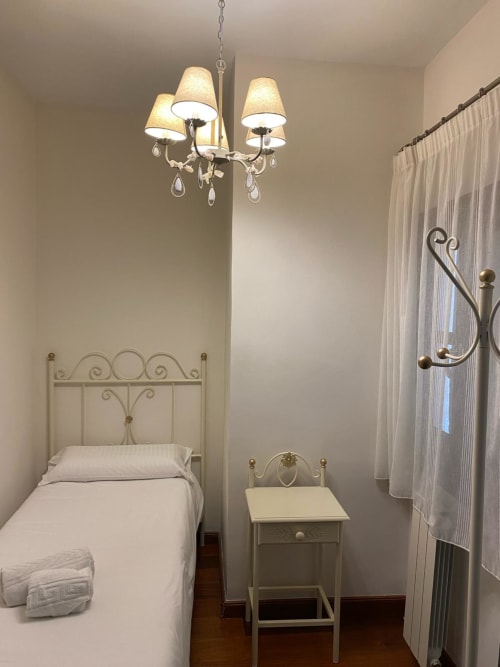 Dormitorio elegante en Suite Comares Granada con cama de metal ornamentada, araña, mesita de noche y cortinas translúcidas