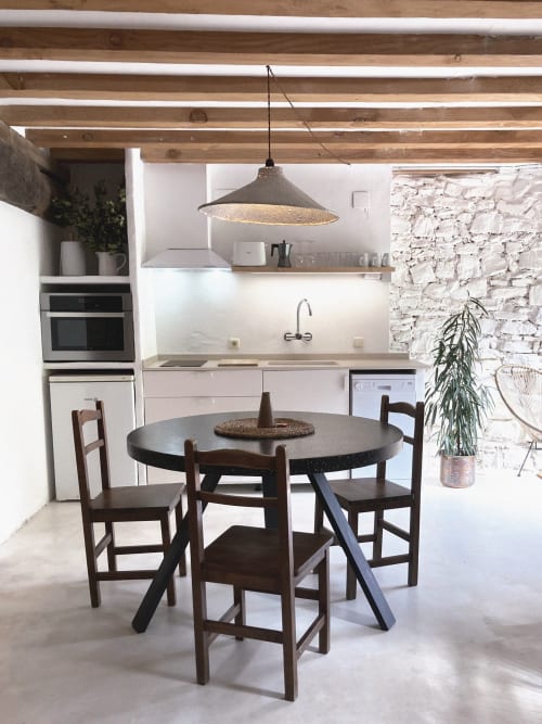 Cocina-comedor rústica moderna con mesa redonda negra, sillas de madera, vigas expuestas, pared de ladrillo blanco y lámpara colgante