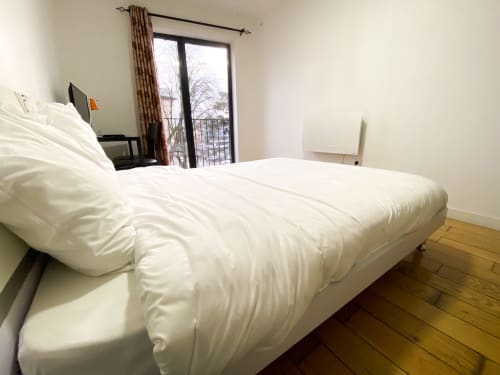 Chambre lumineuse à la Chambre Pasteur à Montreuil : lit blanc, parquet, bureau et porte-fenêtre vitrée avec rideaux