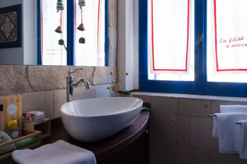 Bagno moderno del Le Vajane B&B con lavabo ovale bianco, ripiano in legno scuro, rivestimento in pietra e finestre con vista mare e dettagli blu