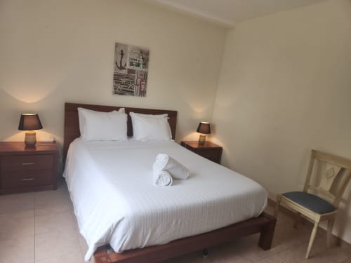 Cama de casal com lençóis brancos, toalhas enroladas, mesas de cabeceira com luminárias e cadeira em quarto claro da pousada Sunny House, Lagos