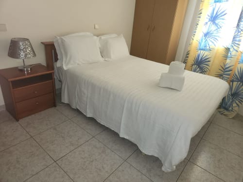 Lit double aux draps blancs dans une chambre de l'auberge Sunny House à Lagos, avec table de nuit en bois, lampe et plante bleue