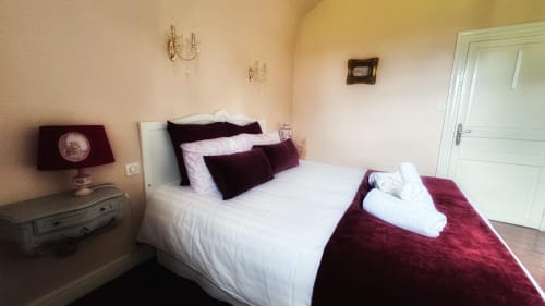 Chambre élégante du Manoir Le Coutureau avec lit blanc, oreillers bordeaux, lampe de chevet et porte vers la salle de bains