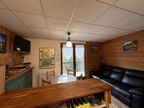 Séjour avec lambris bois, table à manger, canapé cuir noir, kitchenette et portes-fenêtres aux Gîtes Des