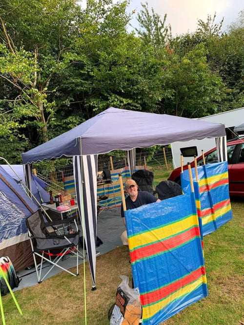 Camping setup at Postwood Gardens Campsite with canopy tent, chairs, and colorful striped storage in Kentisbeare