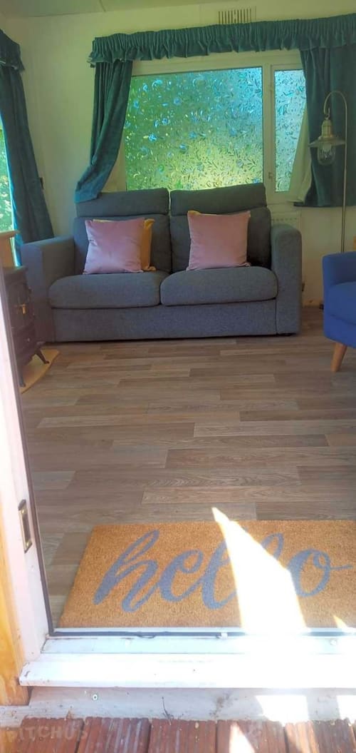 Welcome mat at entrance of Postwood Gardens Campsite guest lounge with grey sofa and green curtains in Kentisbeare, GB