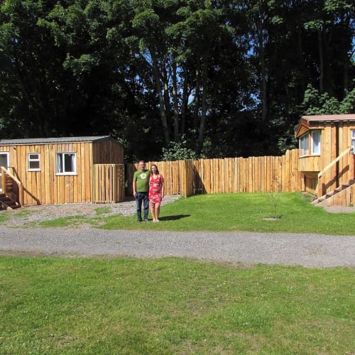 Couple standing in grassy courtyard at Postwood Gardens Campsite in Kentisbeare with wooden cabins and trees