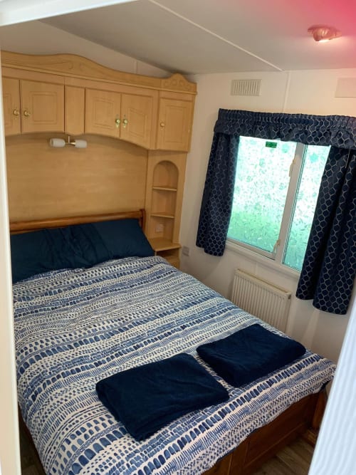 Cozy bedroom in Postwood Gardens Campsite caravan with wooden furniture, patterned bedding, and window with curtains