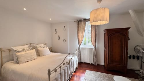 Chambre lumineuse du Manoir Le Coutureau avec lit en métal, lampe suspendue, armoire en bois foncé et fenêtre sur le jardin