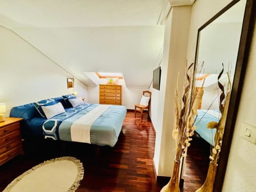 Amplio dormitorio en buhardilla del apartamento de Baiona con ropa de cama azul, muebles de madera, tragaluz y suelos de madera