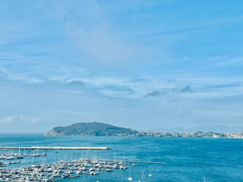 Marina de Baiona con veleros, bahía turquesa y pueblo costero con isla boscosa de fondo