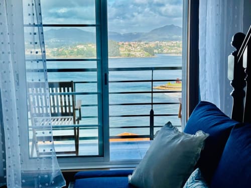 Terraza de apartamento en Baiona con vistas al mar y montañas, cortinas blancas, cojines azules y vistas del pueblo costero