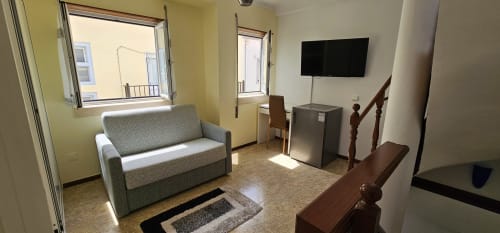 Sala de estar do apartamento Sunao Nazaré com sofá cinzento, televisão de ecrã plano, janelas e corrimão de escada em madeira