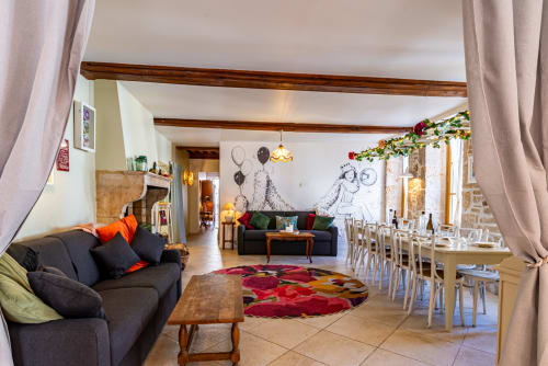 Open-plan living area at La Maison George with dark sofa, colorful rug, dining table, and wooden beams