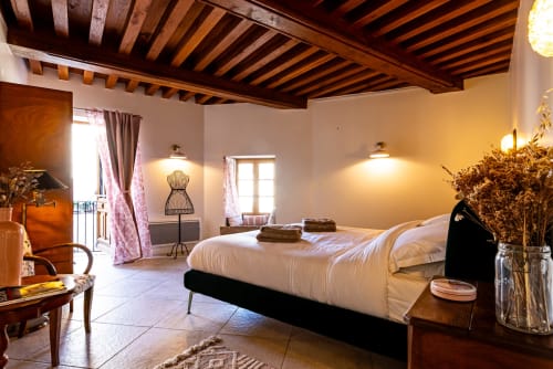 Spacious bedroom at La Maison George with exposed wooden beams, white bed, elegant furnishings, and balcony access in
