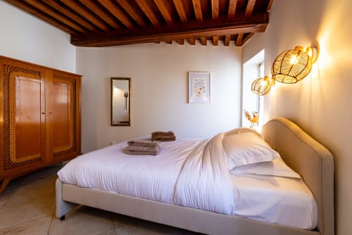 Elegant bedroom at La Maison George with exposed wooden beams, cream bed, wooden wardrobe, and ambient gold pendant lighting