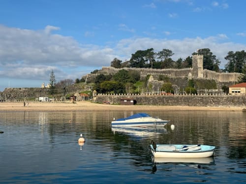 Paseo marítimo de Baiona con murallas medievales, barcos de pesca amarrados y playa arenosa bajo cielo azul