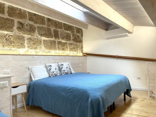 Modern loft bedroom at Catalogna10 with blue quilted bed, exposed stone wall, wooden beams, and city views in Alghero