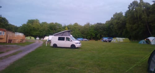 White camper van with pop-up roof parked on grassy field at Postwood Gardens Campsite, Kentisbeare, surrounded by trees and