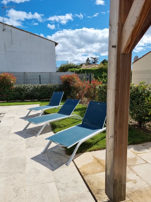 Sunny patio with three blue lounge chairs at La Conciergerie De Franck villa in Boissieres, bordered by flowering plants