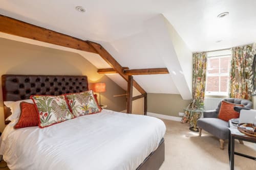 Bedroom 3 in the bed and breakfast; queen size bed with leather headboard, grey upholstered armchair, wooden side table with coffee maker, and old ceiling beams