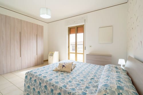 Camera da letto luminosa fronte mare presso Lux Immobiliare con biancheria azzurra a fantasia, armadio in legno e balcone vista Marina di