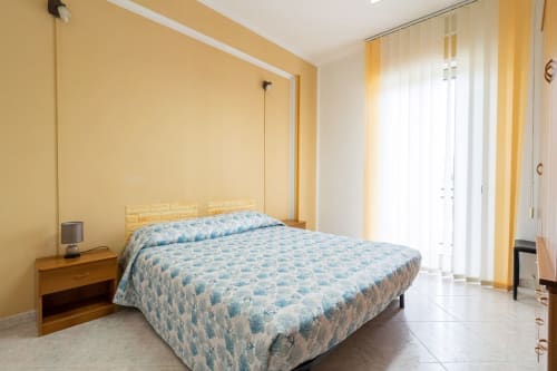 Camera luminosa fronte mare con biancheria blu fantasia, pareti gialle, comodino in legno e balcone vista mare a Marina di