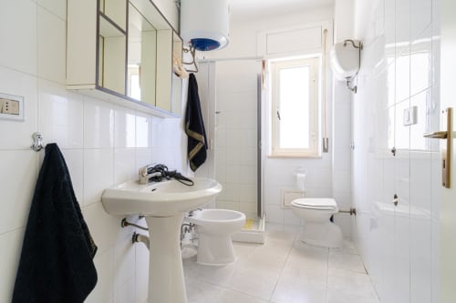 Bagno moderno con piastrelle bianche, lavabo su piedistallo, wc e doccia in vetro nell'appartamento fronte mare di Lux Immobiliare a Marina