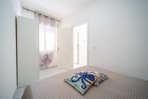 Camera luminosa con cuscini decorativi su letto trapuntato, specchio e porte che si aprono sul balcone con vista mare da Lux Immobiliare