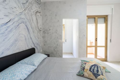 Camera luminosa con parete in marmo, letto matrimoniale, biancheria blu, porte in legno con vista sul mare a Lux Immobiliare
