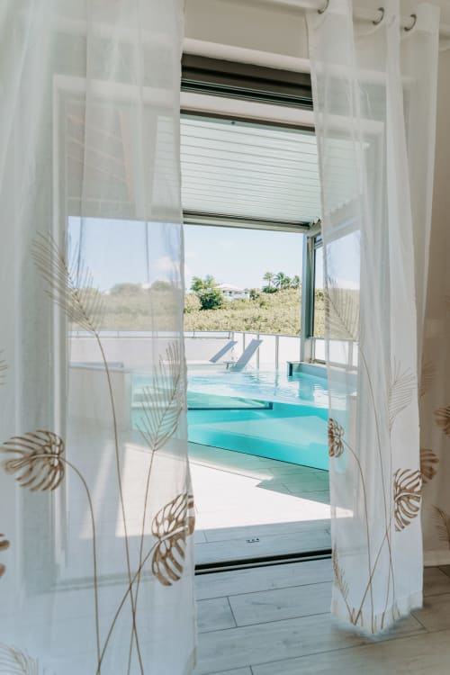 Des rideaux voilages à motifs de feuilles de palmier encadrent la piscine turquoise de l'Habitation l'SEO, Sainte-Anne, Guadeloupe
