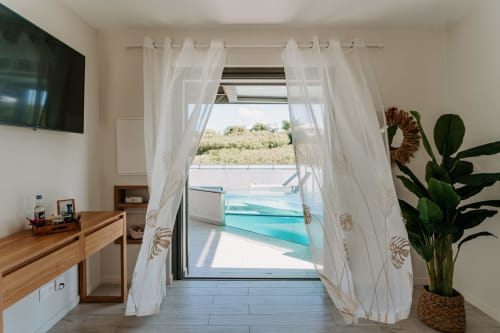 Modern hotel room with sheer curtains framing glass doors opening to turquoise pool and garden at Habitation I'SEO,