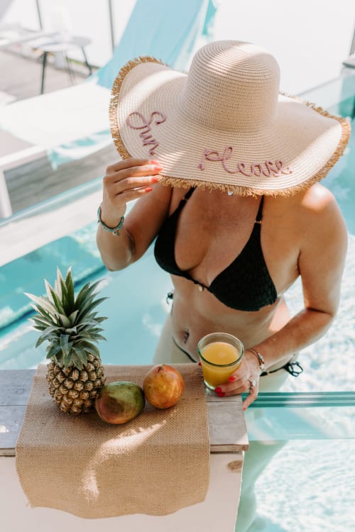 Femme en bikini noir au bord de la piscine d'Habitation l'SEO, Sainte-Anne, tenant un chapeau à large bord avec fruits tropicaux et jus