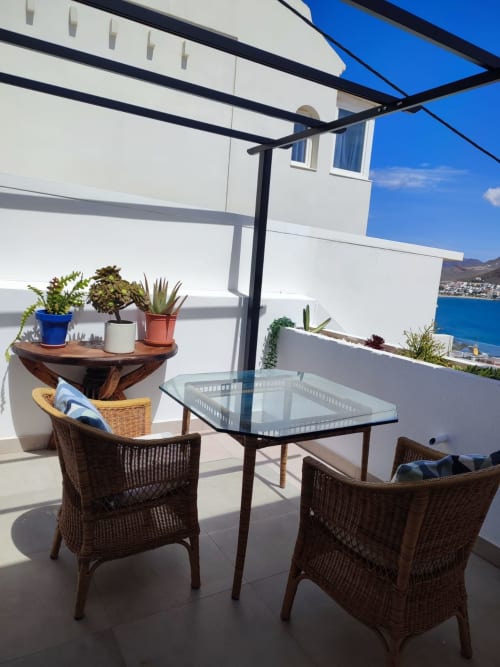 Terraza soleada en El Faro De Los Genoveses con sillas de mimbre, mesa de cristal, vistas al mar de San José, Almería, y sombrilla azul