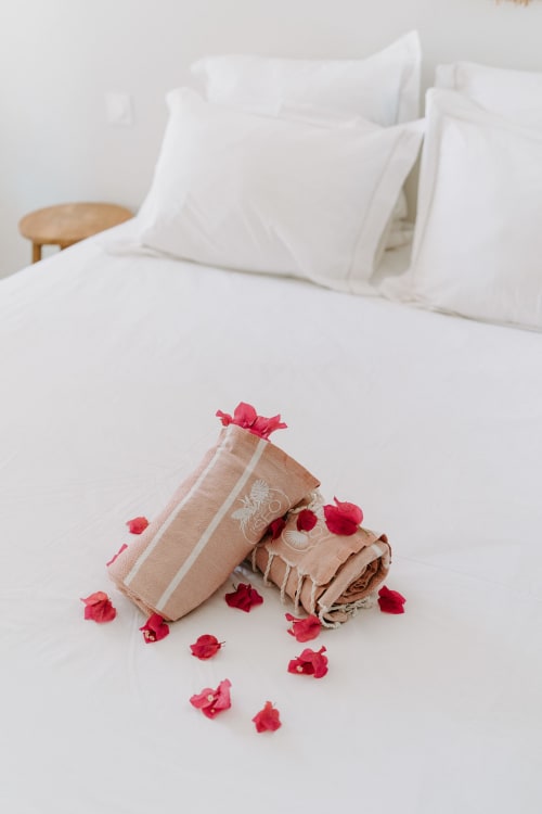 Chambre luxueuse avec oreillers et draps blancs, serviettes beige nouées de ruban rouge et pétales de rose à l'Habitation I'SEO
