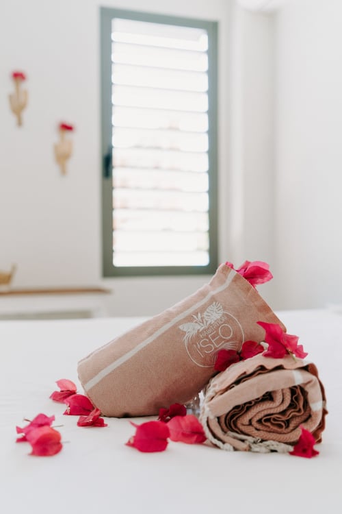 Équipements spa à Habitation l'SEO : serviette beige au logo, pétales de rose et tissu roulé dans une salle de bain lumineuse avec volets