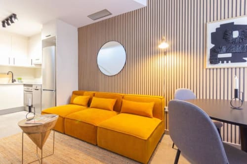 Estudio moderno J&A Apartments en Terrassa con sofá amarillo, pared de acento de madera, espejo circular y sala de estar abierta.