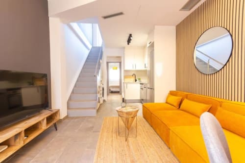 Sala de estar de los Apartamentos J&A en Terrassa con sofá amarillo brillante, mueble TV de madera, escaleras y espejo redondo