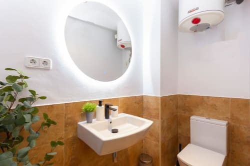 Baño moderno en los Apartamentos J&A en Terrassa con espejo iluminado, lavabo blanco, inodoro y detalles de azulejos de madera cálida