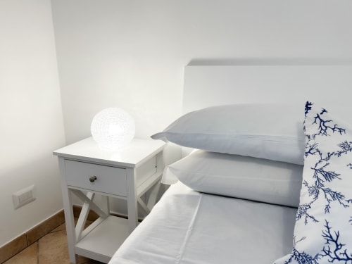 Chambre minimaliste à CefalHome Roma avec table de nuit blanche, lampe texturée et coussins bleus à motifs