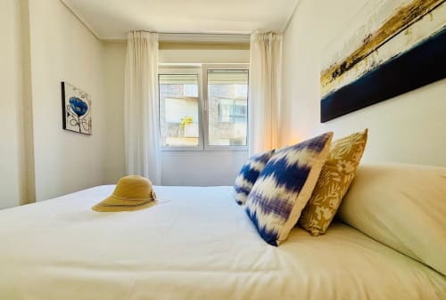 Dormitorio luminoso en apartamento Breixo, Baiona con ropa de cama color crema, almohadas azules y doradas, sombrero amarillo y vistas a la ciudad
