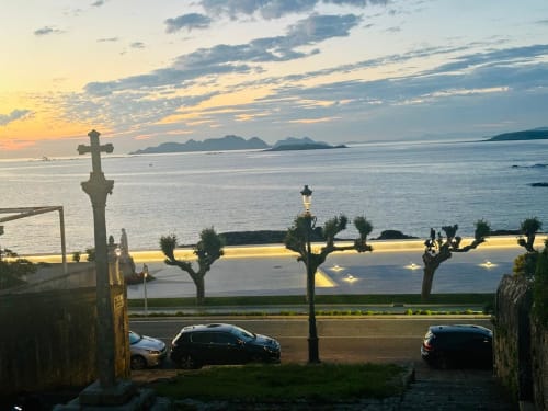 Vista al atardecer sobre la bahía de Baiona desde el apartamento Breixo, con cruz celta y coches aparcados, con vistas a las montañas y la costa