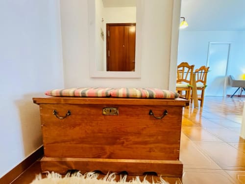 Baúl de madera con cojín rayado en apartamento de Baiona con vistas al mar, sillas de madera visibles en salón luminoso