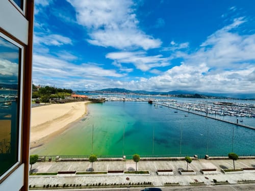 Vista del apartamento frente a playa de Baiona: playa de arena, bahía turquesa, puerto deportivo con embarcaciones, montañas y cielo azul despejado