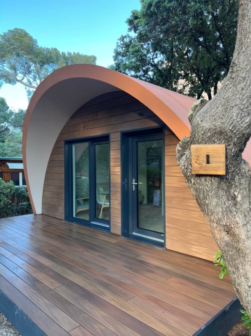 Cabine pod moderne aux lignes courbes à Camping La Lézardière avec terrasse bois, portes vitrées et toit en terre cuite parmi les arbres